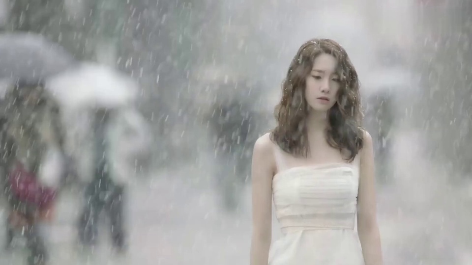 雨天街景中，穿米白色抹胸礼服的棕发女性低头站立，神情低落，背景虚化朦胧，左侧有撑伞行人的模糊轮廓。