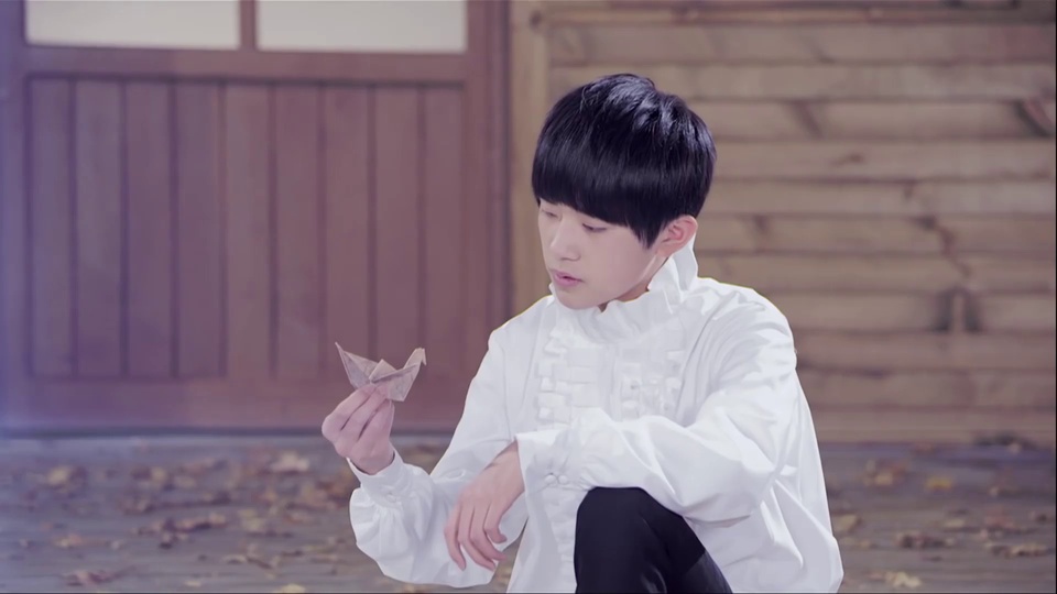 tfboys主题真人剧照里，身着白衬衫黑长裤的短发年轻男子站在木门旁的木地板上，手边放着一只浅米色千纸鹤，地面散落黄褐色枯叶，整体带有淡紫色柔化滤镜色调。