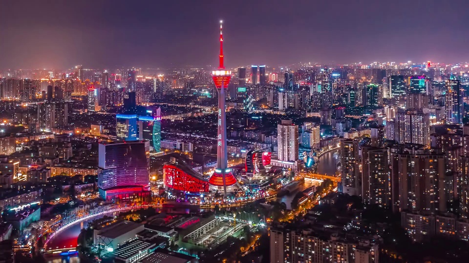 高空俯瞰的城市夜景，中心矗立带红色观光层的电视塔，河道穿插亮灯的城市建筑群