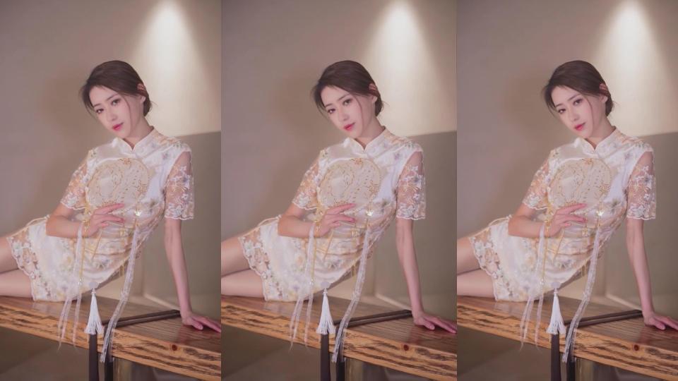 穿浅杏色蕾丝短旗袍的女子手持流苏团扇倚靠木质桌沿，暖光氛围柔和雅致。