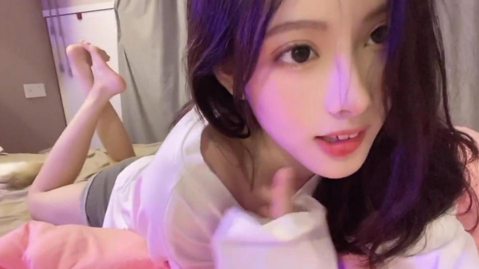 紫粉柔光笼罩的室内床铺上，深棕长发女生趴卧对着镜头，双腿向上翘起。