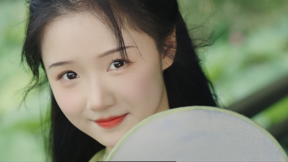 持白色半透明团扇的黑长发古风女子，背景为虚化的浅绿色户外植物环境。
