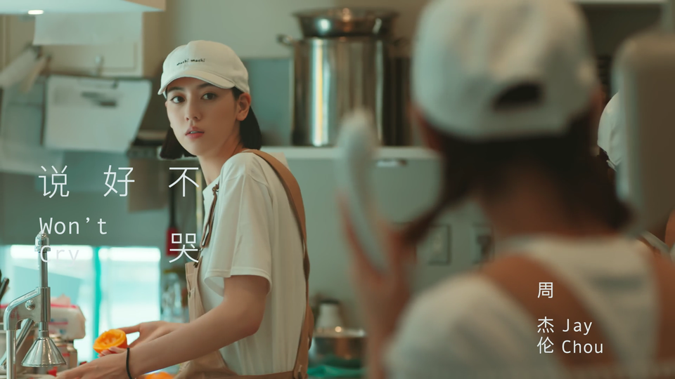 写实风格的奶茶店后厨内，黑色齐耳短发的女店员头戴印小字的白色棒球帽，身着白短袖与卡其色背带围裙，手持半块橙子，前景有模糊的同款着装店员背影，背景摆放不锈钢容器与厨房电器。