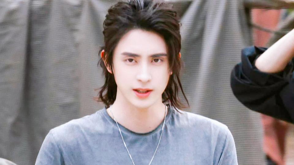 留深棕微卷长发、穿灰蓝色圆领T恤的敖瑞鹏站在灰色幕布前的户外片场场景。