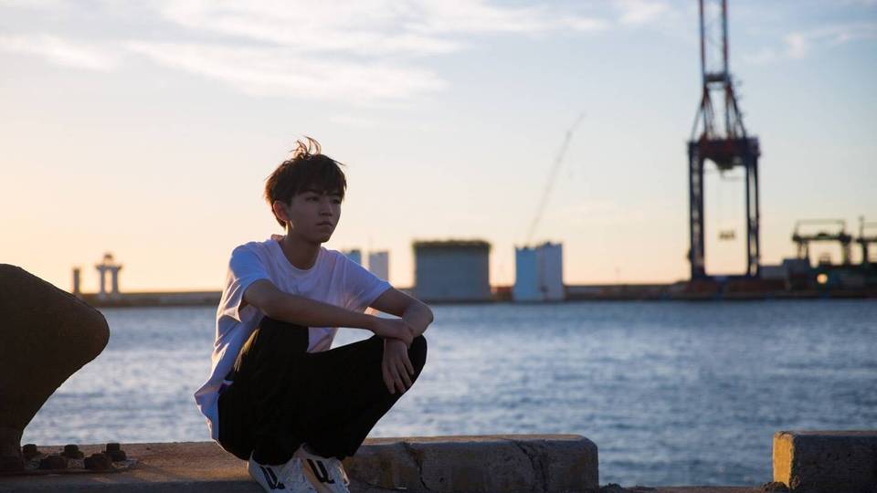 穿白短袖黑长裤的年轻男性蹲在临海码头混凝土台面上，望向画面右侧，背景是黄昏渐变天空下的蓝色海面与港口起重机、低矮仓储建筑。