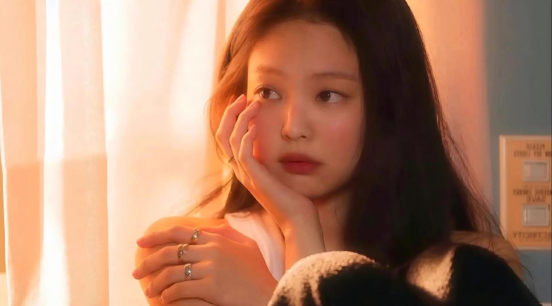 暖橙色夕阳光线下，jennie单手托腮坐在室内窗边，旁侧有透光纱帘与蓝色墙面，佩戴多枚银戒指，身穿带白色毛绒边的上衣。