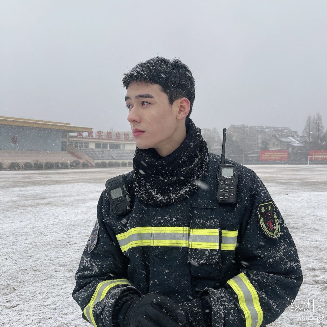飘雪训练场中身着带反光条黑色消防制服的短发男性，制服落有白雪，背景可见覆雪场地与远处的训练场馆建筑