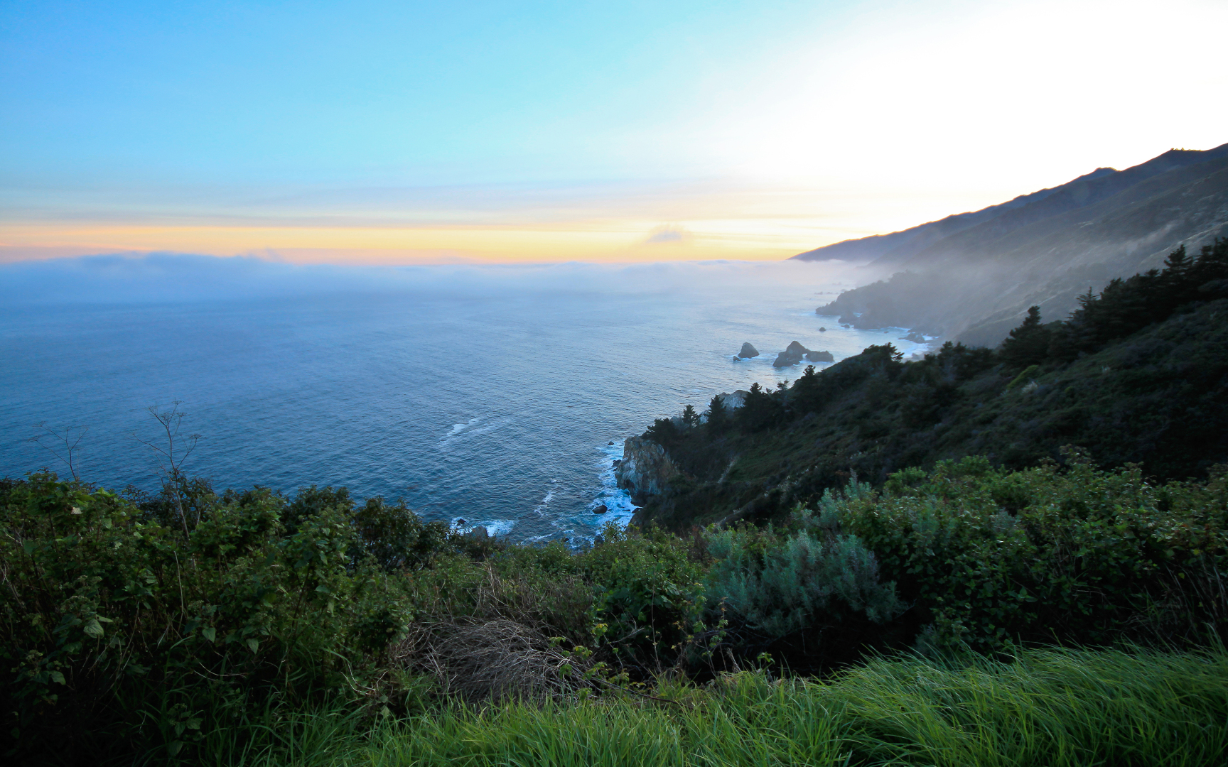 写实风格海岸日落风景，薄雾笼罩的翠绿层叠山坡下是蔚蓝开阔海面，海天交界晕着橙蓝渐变的黄昏天色，前景分布层次丰富的自然植被。