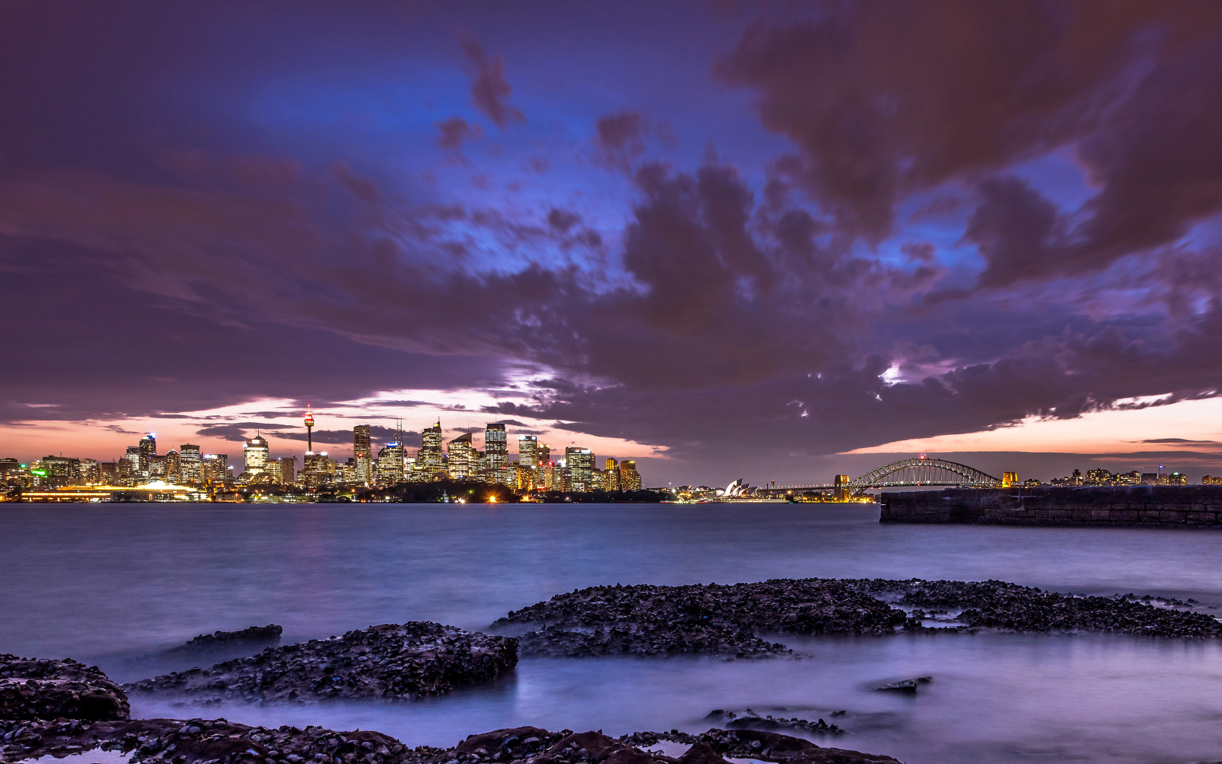 写实摄影画面，紫橙渐变暮色天空下的悉尼港湾，可见海港大桥、悉尼歌剧院与亮灯的CBD天际线，前景是平静海湾与露出水面的礁石。