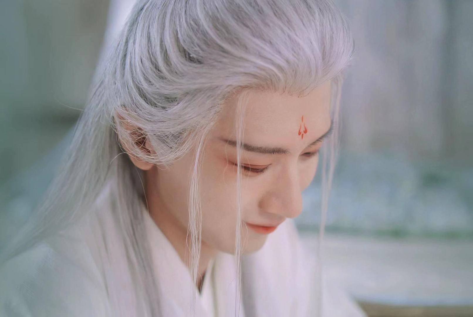 成毅古风形象照，银白长发配白色古风服饰，额间红印，背景为浅青绿色虚化朦胧户外场景