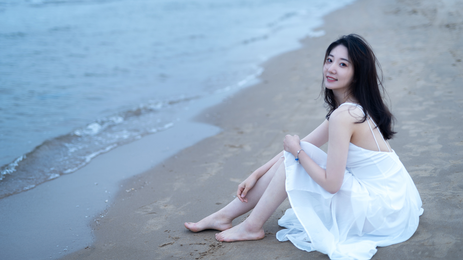 穿白色露背吊带长裙的年轻女性坐在海边浅沙色沙滩上，赤足朝向镜头微笑，身后是漫着柔浪的浅蓝色近海。