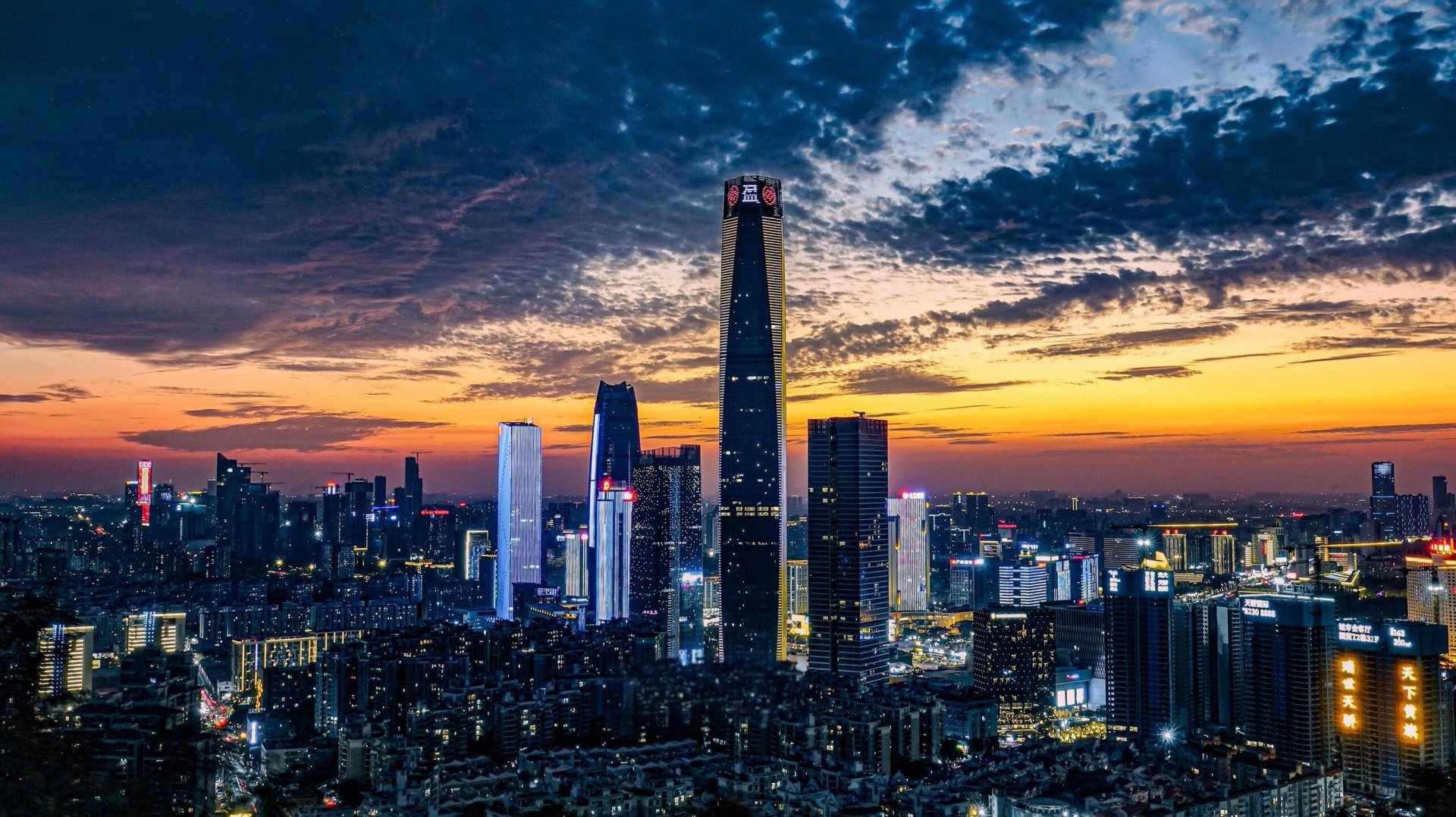 黄昏高空视角城市天际线，橙紫渐变晚霞下，连片高楼亮起各色城市灯光。