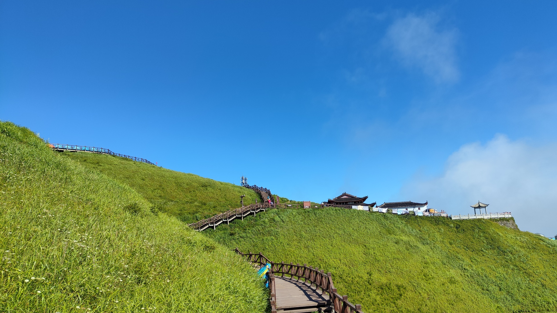 绿草丛生的缓坡山顶，木栈道蜿蜒向上，坡顶有中式古建与凉亭，上方是湛蓝晴空。