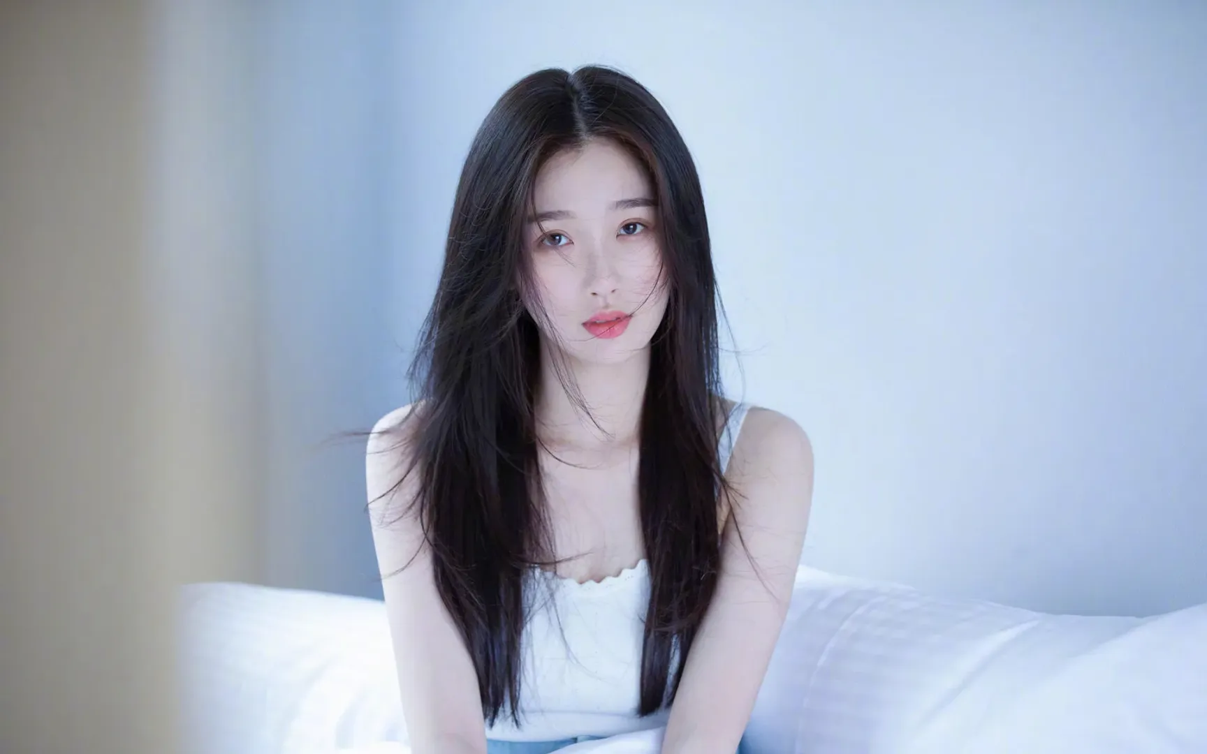身着浅蓝白吊带睡裙的黑色长发年轻女性跪坐在铺有蓬松白色床品的床面上，侧脸朝向镜头，为室内写实摄影画面。