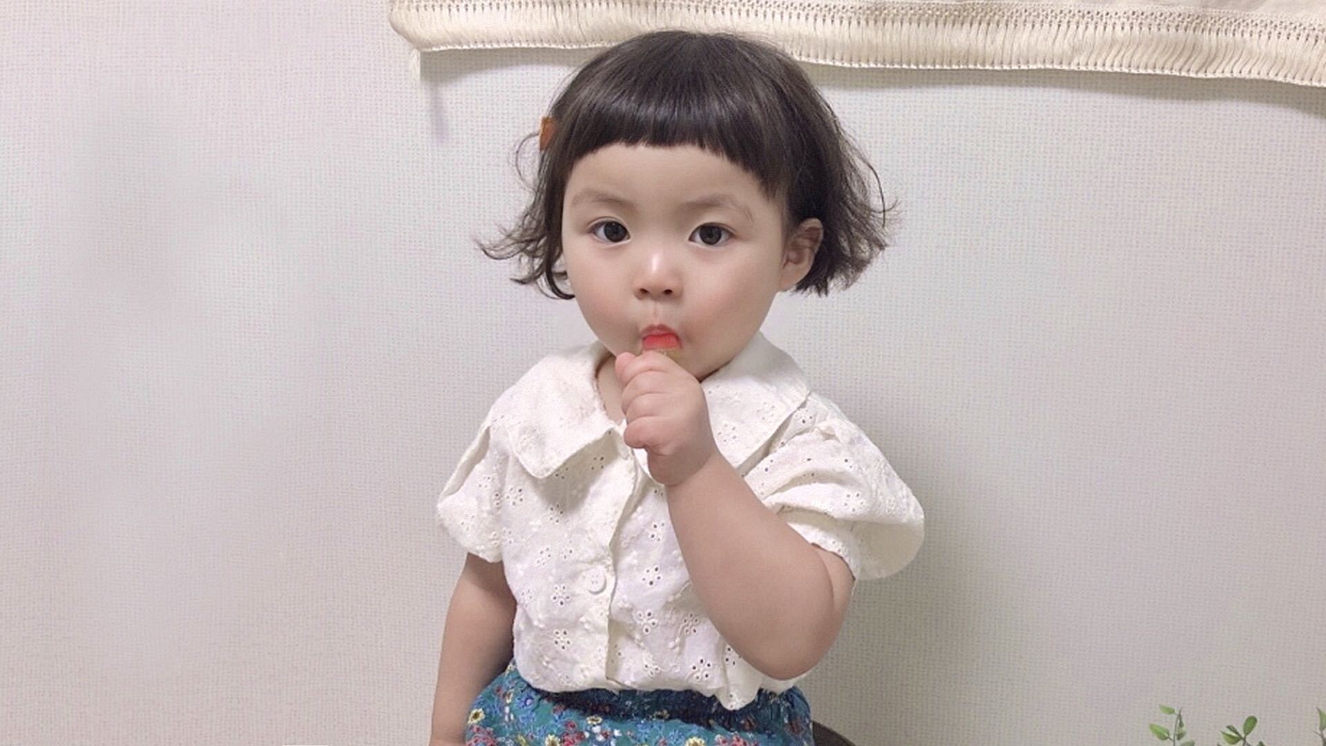 穿白色镂空短袖上衣的短发幼童在浅肌理墙前手持红色小物件