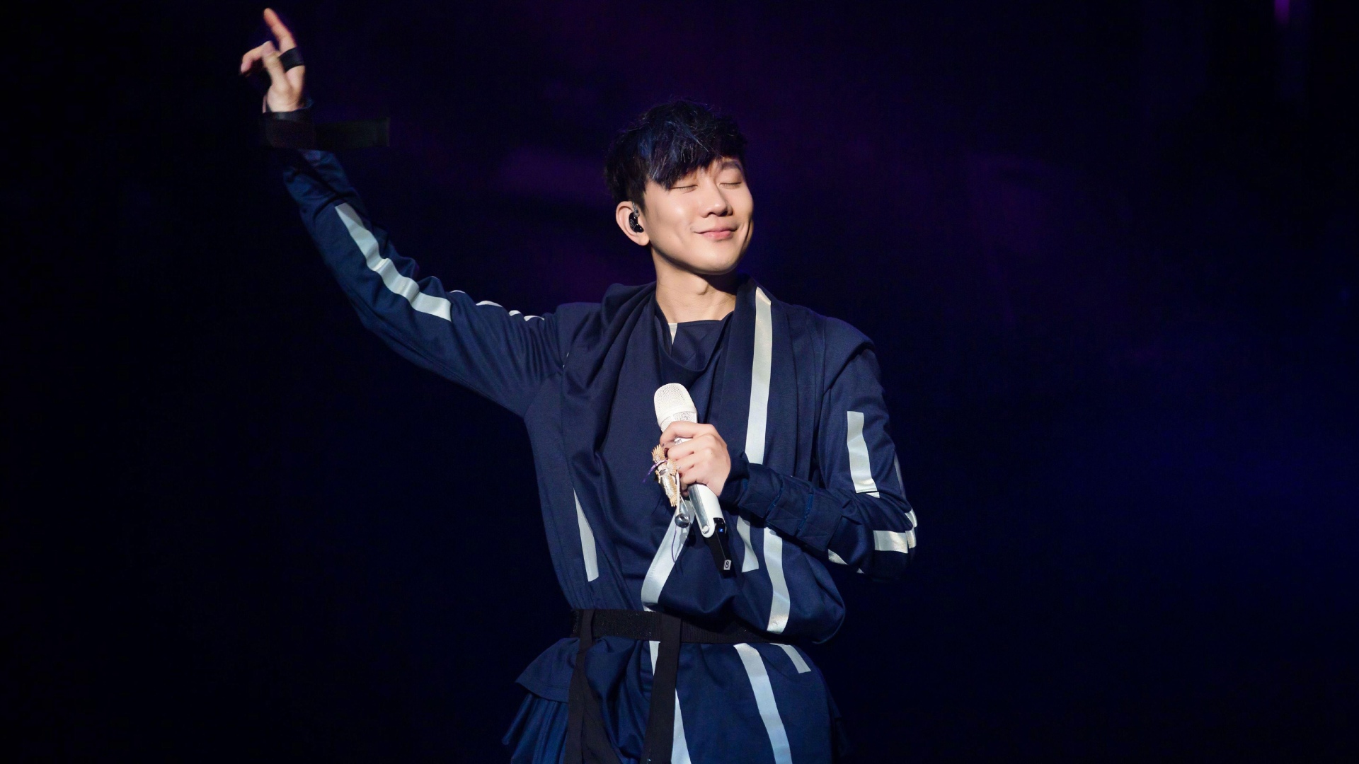 舞台上的年轻东亚男性表演者，穿藏蓝色带银白条纹连体演出服，左手持白色麦克风，右臂抬起比出指向斜上方的手势，双眼闭合面带笑意，背景为深黑搭配暗紫色弥散暗光。