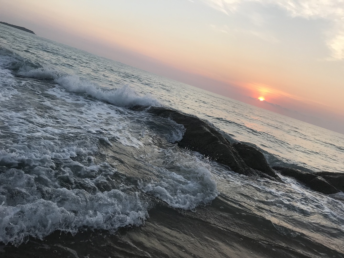 日落时分的海岸，白浪拍击深色礁石，海面延伸至远方，天际晕染粉橙渐变霞光。