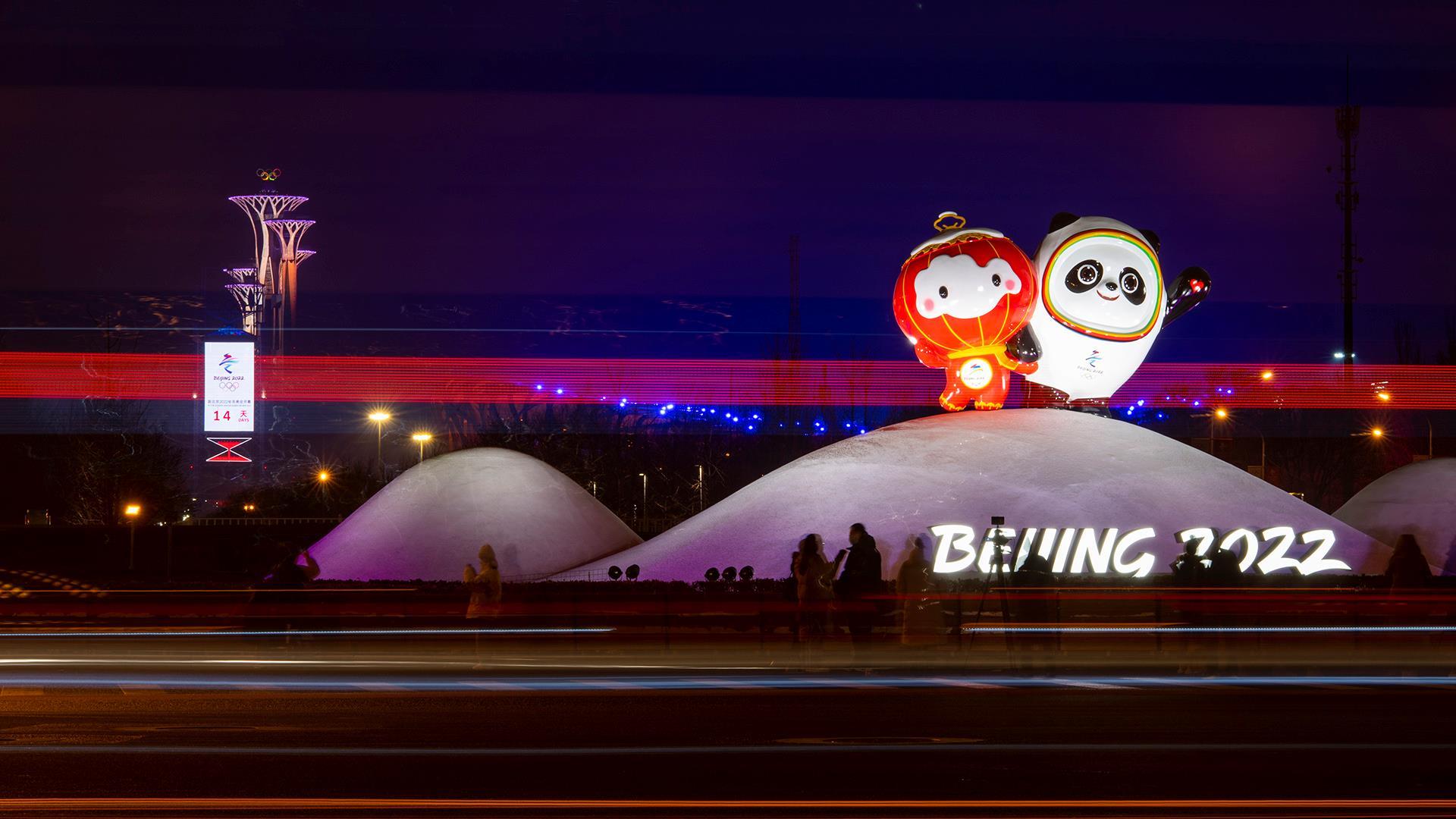 冬夜城市街景，两个北京冬奥吉祥物立在雪白色坡顶装置上，前景是车流拉出的彩色光轨，远处亮着紫灯的奥林匹克塔矗立在暗蓝夜空下。