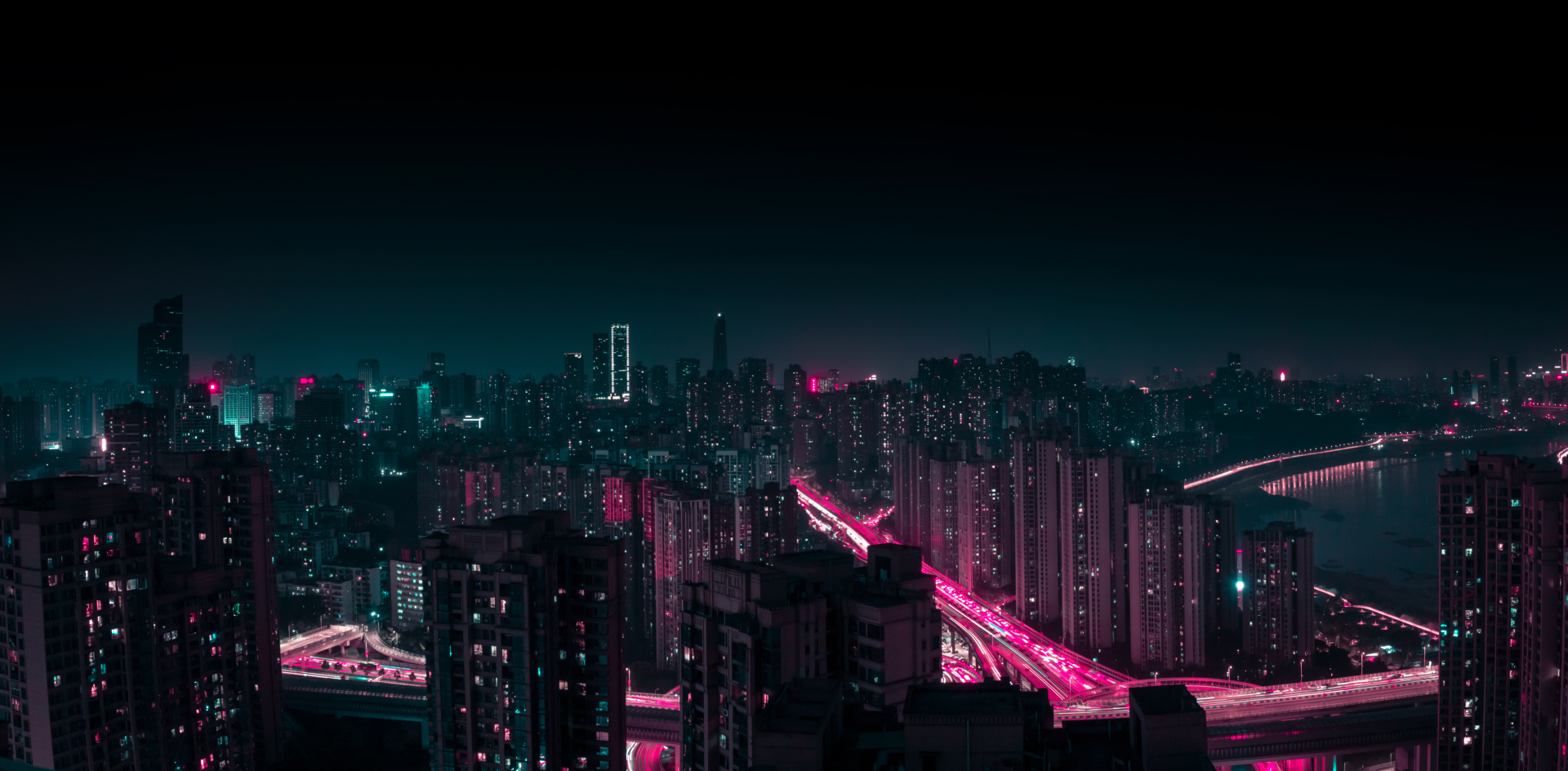 高空视角下的沿江都市夜景，亮着粉青灯光的楼宇连片，高架车流拉出长光轨，江面反射沿岸灯火。