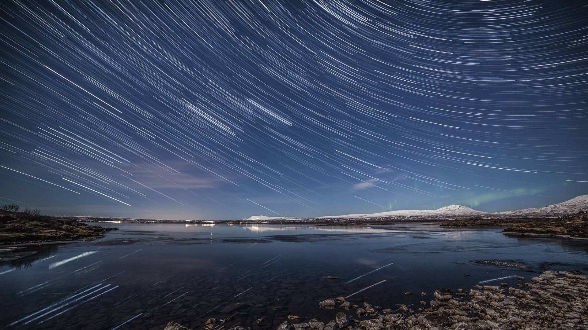 深蓝色夜空布满斜向白亮星轨，湖面倒映星轨，远处是覆雪山峦，岸边分布着碎石与暗色裸地，冷调写实夜景。