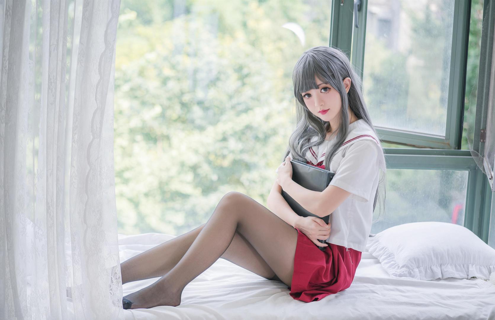 窗边床铺上抱书的水手服少女写真画面。