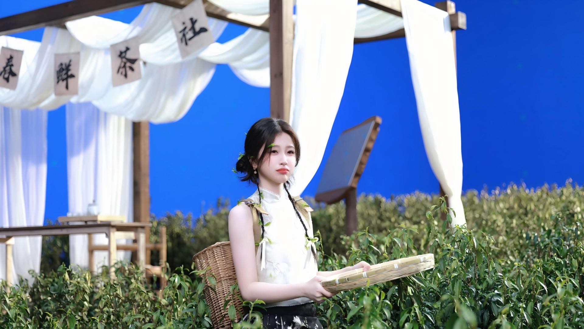梳盘发垂双辫的年轻女性身着白色无袖上衣，发间点缀绿叶，背负竹编背篓，手持竹托盘站在连片翠绿茶丛前，身后立有印着“春 鲜 茶 社”的米色布幡，旁设木质茶寮，背景为纯亮蓝色。